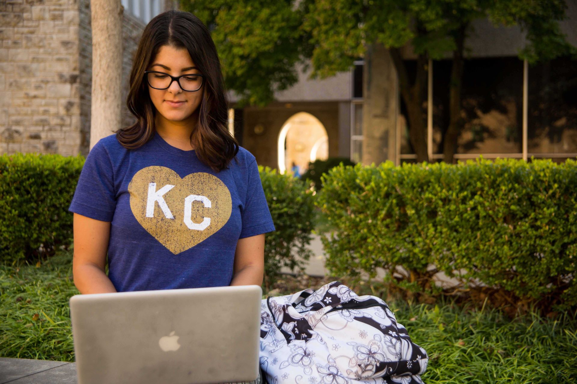 A student stands at a table outside working on a laptop with a backpack beside it